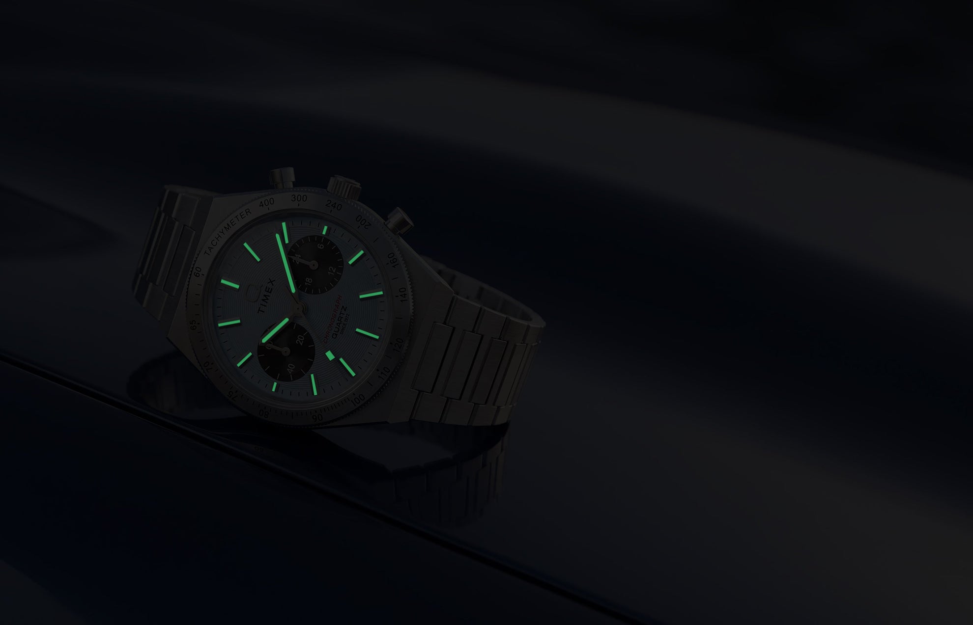 Q Timex Continental Chronograph in low light with green luminous hands and hour markers glowing against a dark blue background.