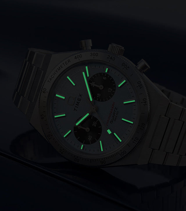 Q Timex Continental Chronograph in low light with green luminous hands and hour markers glowing against a dark blue background.