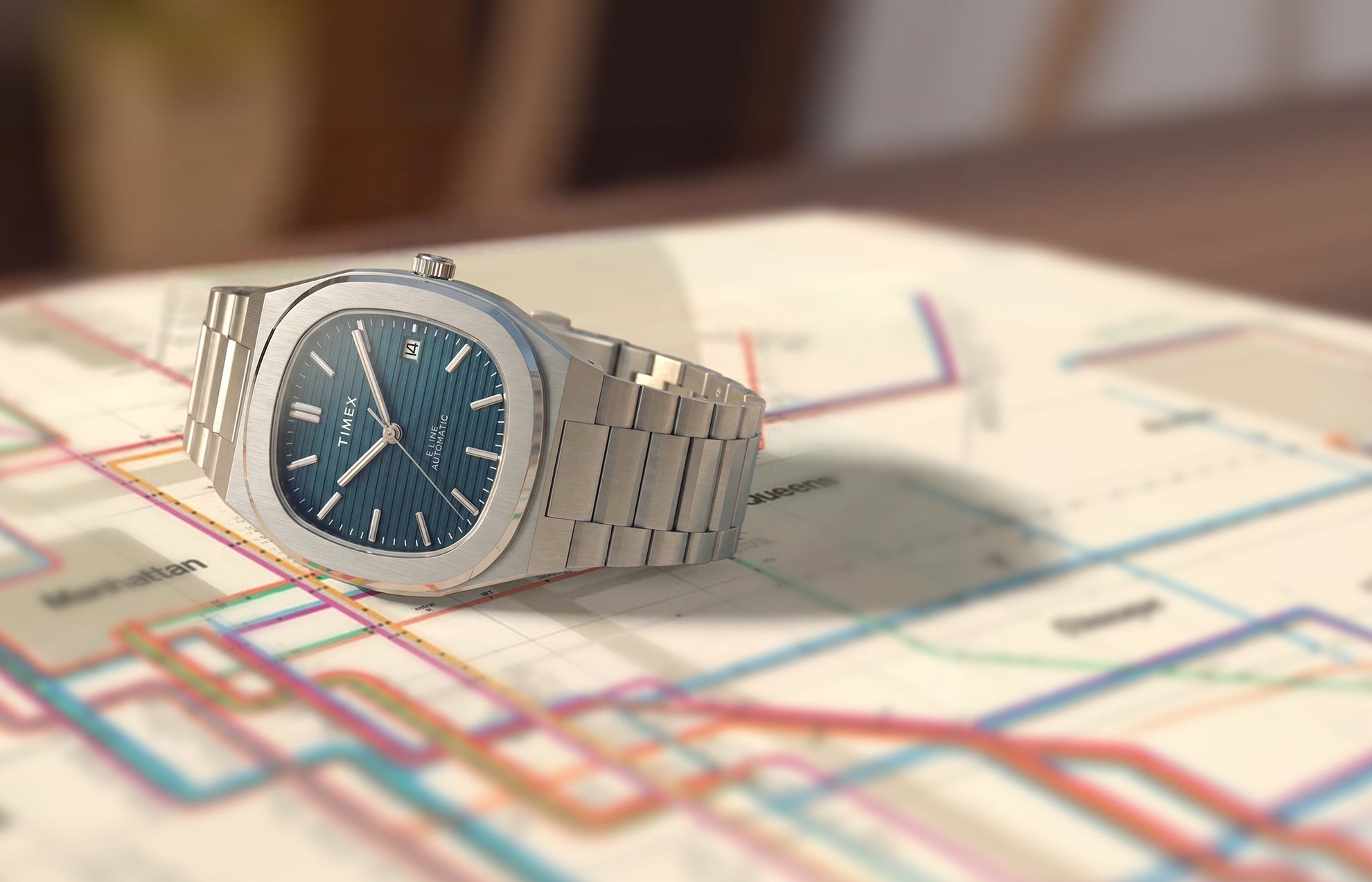 Timex® Automatic E Line 38mm watch with a stainless steel bracelet, blue textured dial, and date window displayed on a transit map.