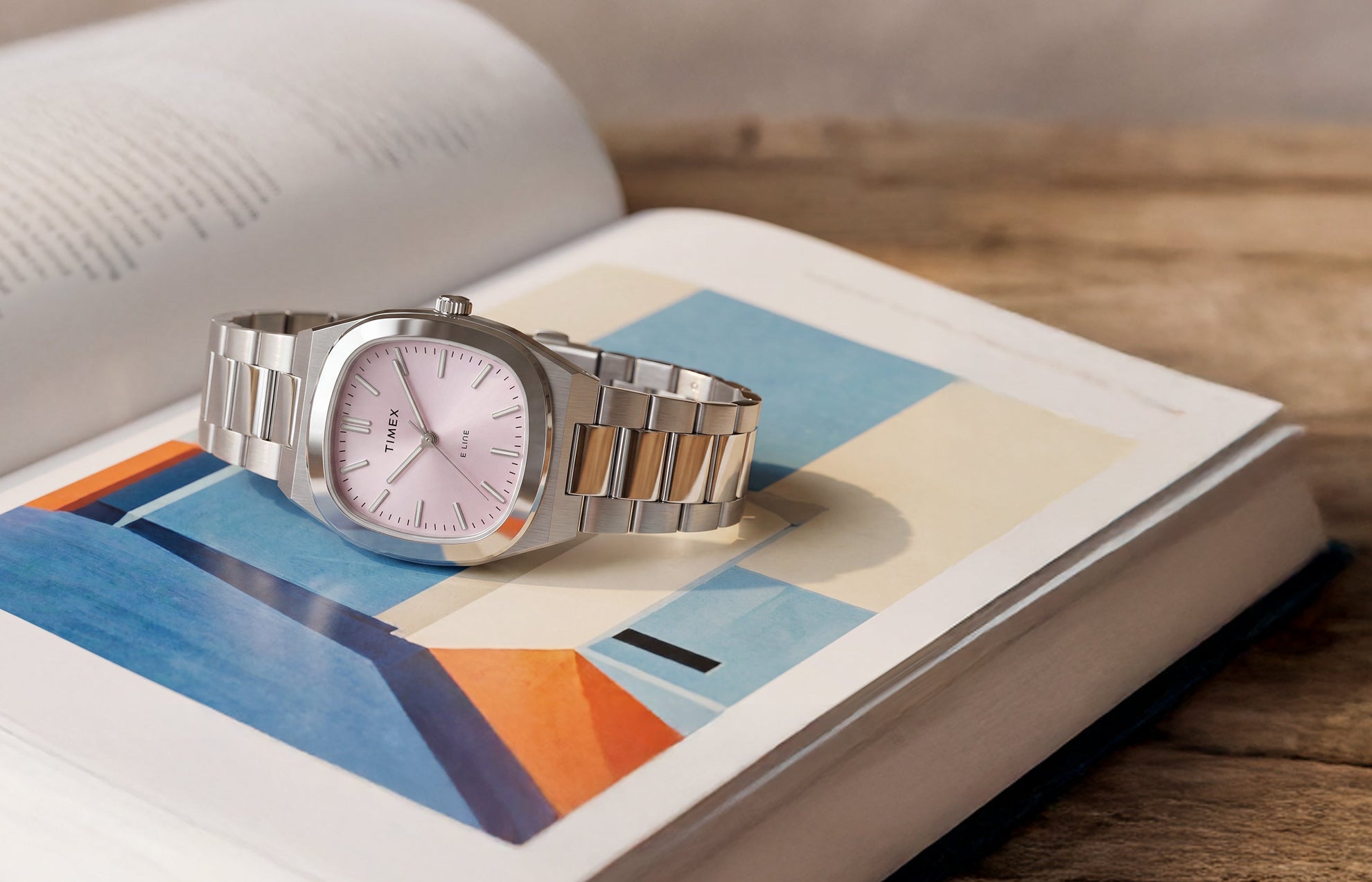 Timex® Quartz E Line 30mm watch with a stainless steel bracelet and pink sunray dial displayed on an open book with colorful artwork.