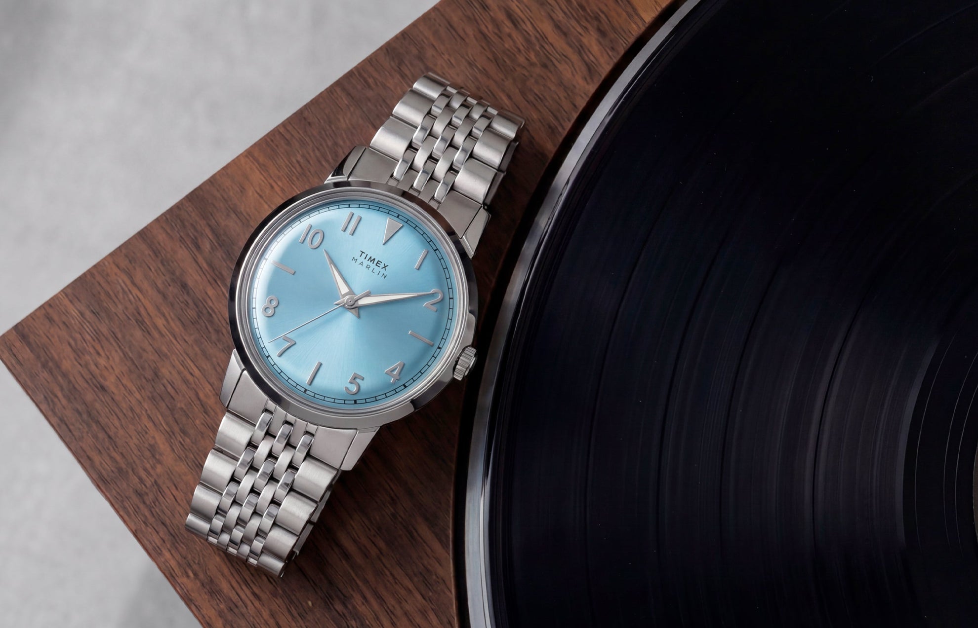 Timex Marlin Quartz watch with an ice blue sunray dial and stainless steel bracelet displayed on a wooden tabletop beside a vinyl record.