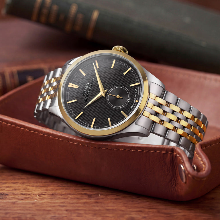 Timex Waterbury Classic watch with a two-tone stainless steel bracelet, gold-tone bezel, black textured dial, and small seconds sub-dial, displayed in a brown leather valet tray.