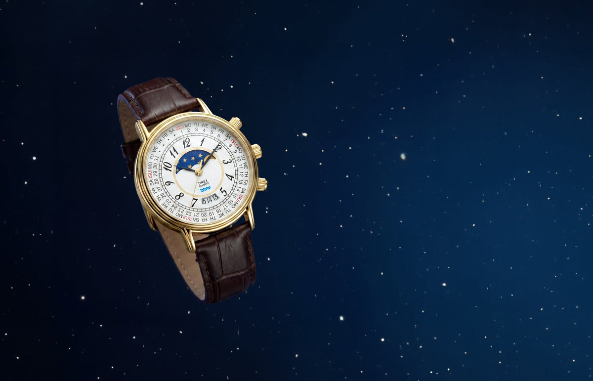 Timex 1990 Moon Phase and Calendar Dial Reissue with a gold-tone case, white calendar dial, moon phase display, and brown leather strap against a starry blue background.
