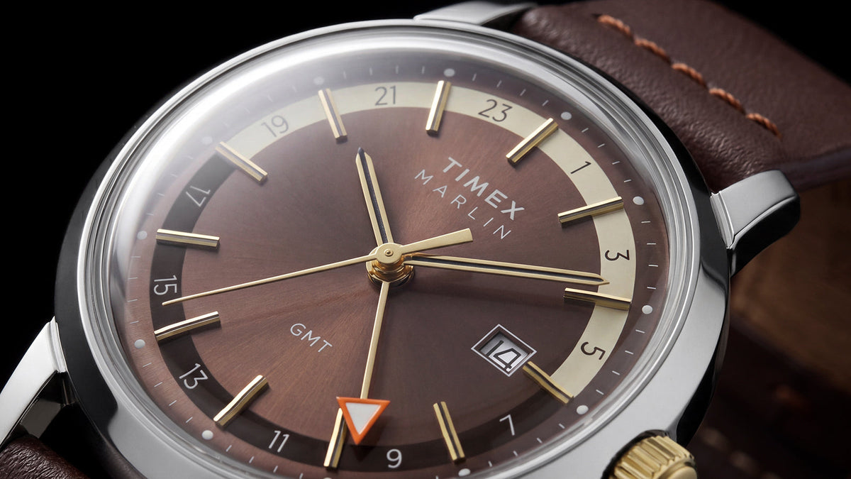 Angled close-up of the Marlin® Quartz GMT dial featuring gold-tone hands, a 24-hour scale, orange GMT hand, and date window.
