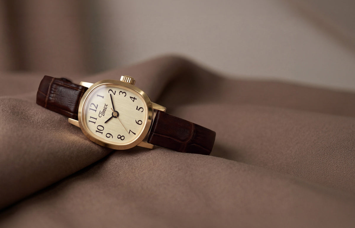 Cavatina® Mini watch with a gold-tone case, champagne dial, and dark brown leather strap resting on soft brown fabric.