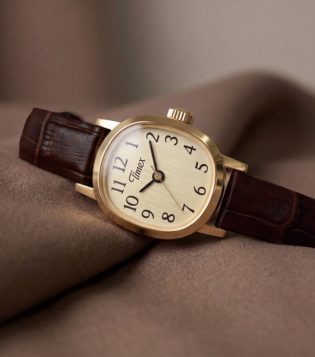 Cavatina® Mini watch with a gold-tone case, champagne dial, and dark brown leather strap resting on soft brown fabric.