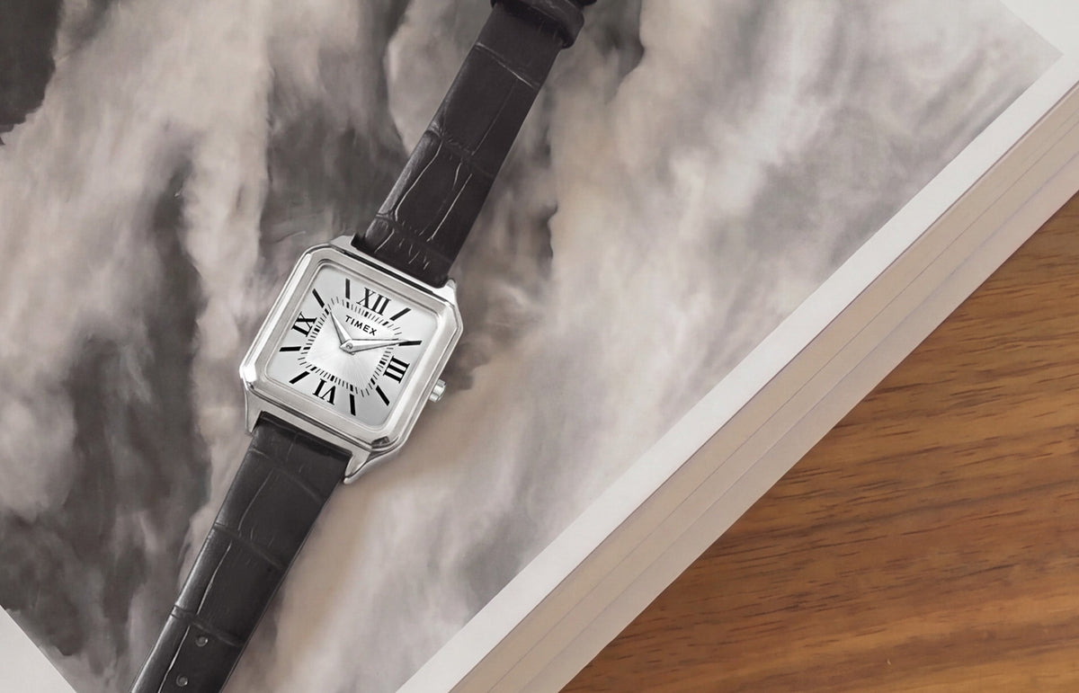 Timex Addison watch with a silver-tone rectangular case, white dial, Roman numerals, and black leather strap displayed on an open book.