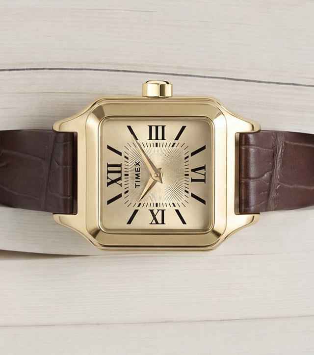 Timex Addison watch with a gold-tone rectangular case, champagne dial, black Roman numerals, and brown leather strap displayed on an open book.