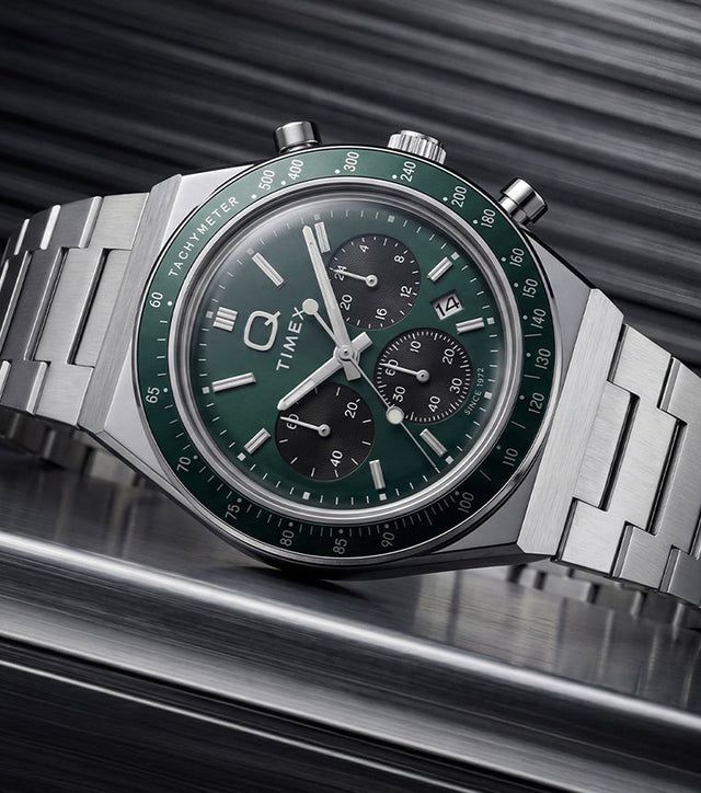 Q Timex Chronograph with a green dial and stainless-steel bracelet displayed on a dark metallic surface.