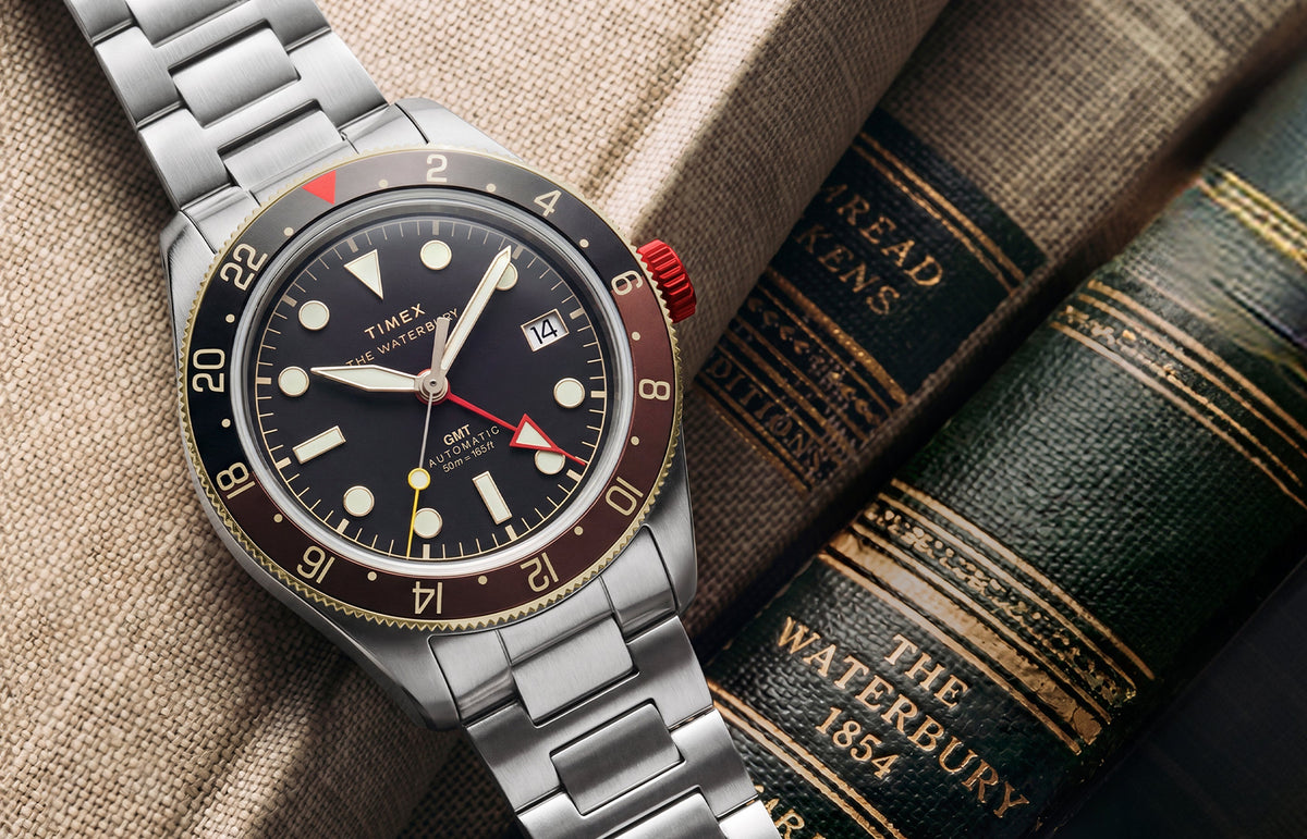 Waterbury Heritage Automatic GMT watch with a black dial, brown 24-hour bezel, red crown, and stainless-steel bracelet displayed beside vintage books.