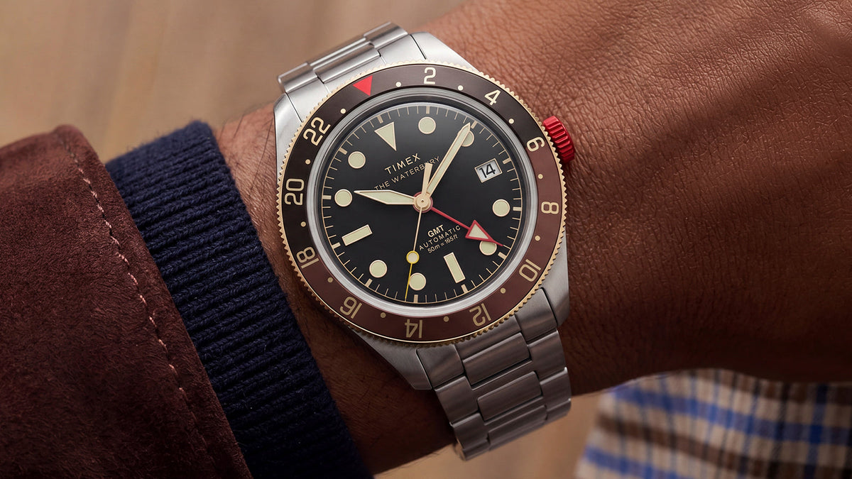 Waterbury Heritage Automatic GMT watch worn on the wrist, featuring a black dial, brown bezel, red crown, and stainless-steel bracelet.