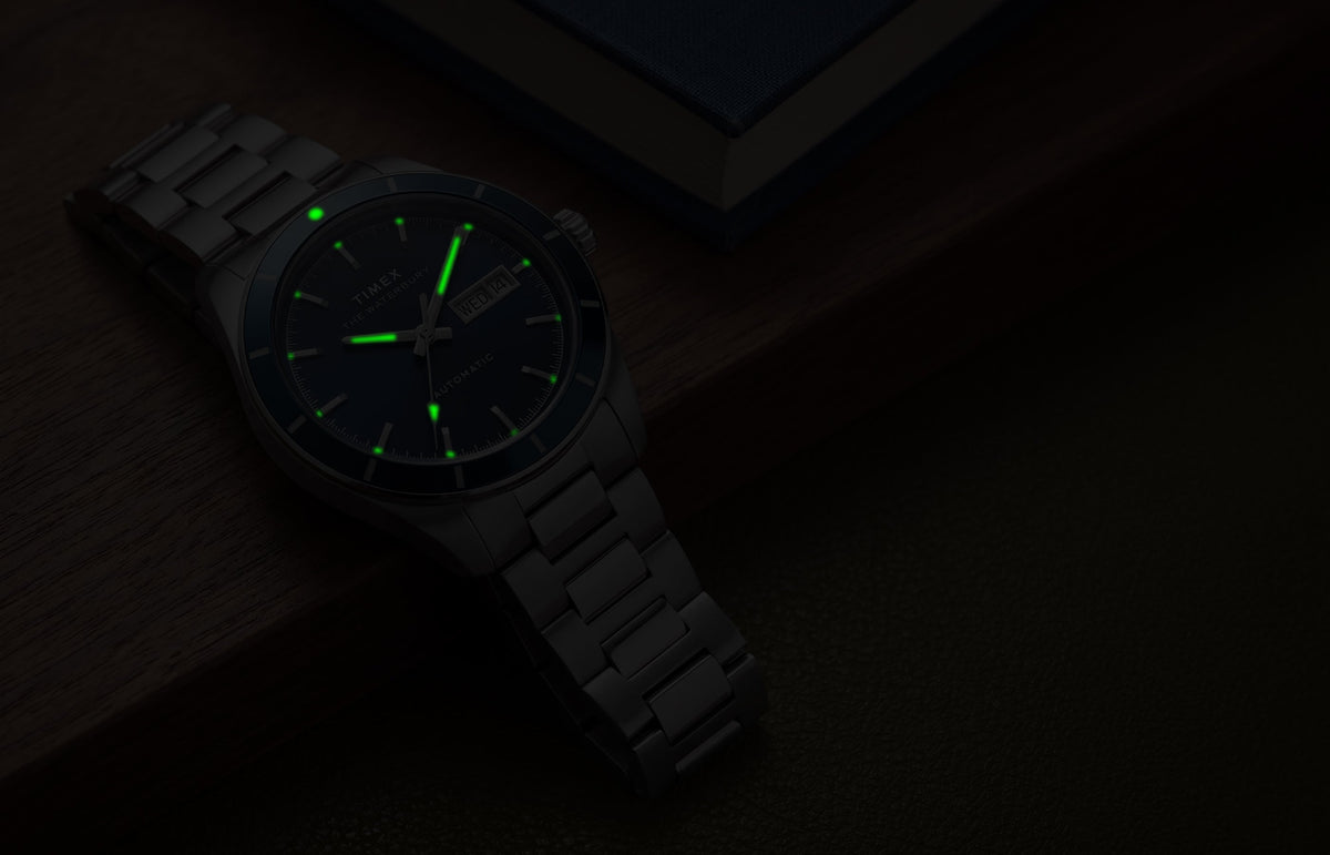 Waterbury Heritage Automatic watch glowing green in low light beside a blue book on a wooden surface.