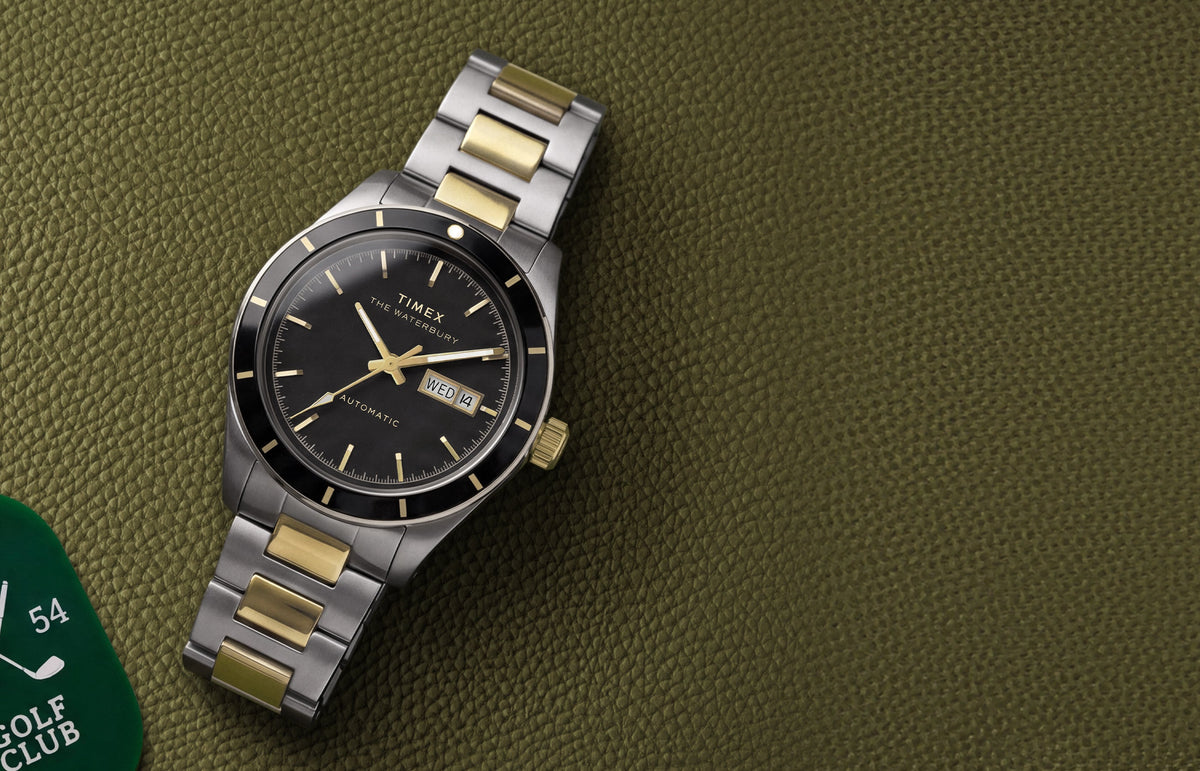 Waterbury Heritage Automatic watch with a black dial and two-tone bracelet resting on textured olive-green leather beside a golf tag.