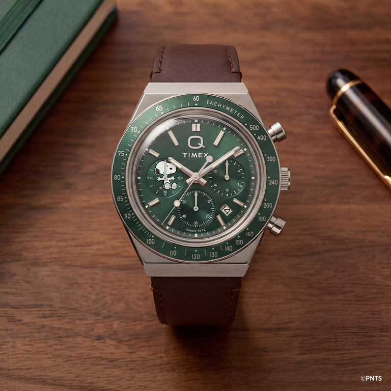 Peanuts x Q Timex Chronograph with a green dial, Snoopy subdial, and brown leather strap displayed on a wooden desk.