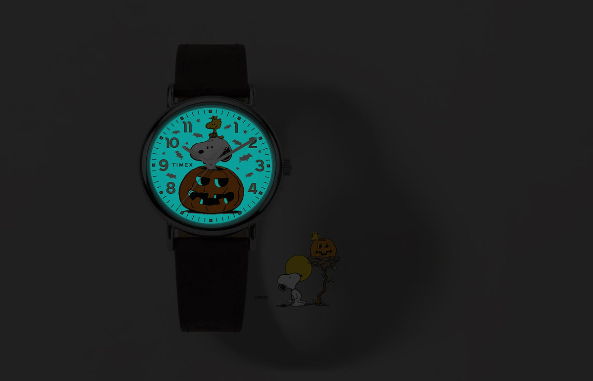 Peanuts® x Timex Weekender® Pumpkin watch shown in the dark with a glowing blue INDIGLO® dial revealing a bat pattern around Snoopy and Woodstock.