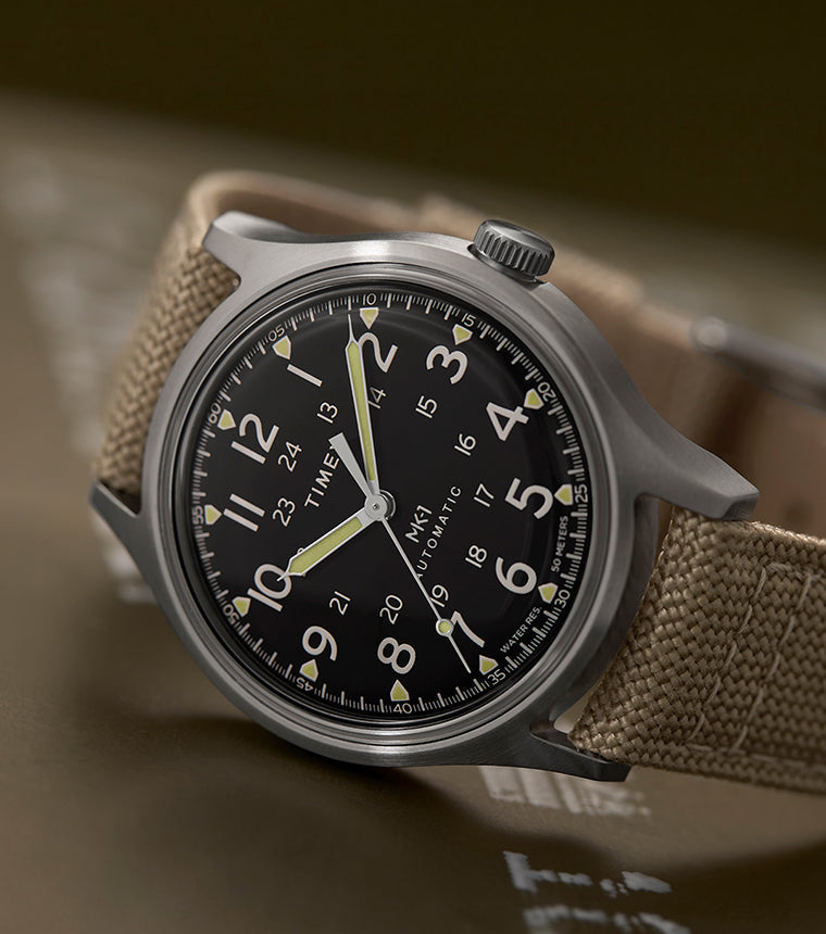 MK1® Automatic watch with a black dial, brushed silver-tone case, and tan fabric strap resting on military-style equipment.