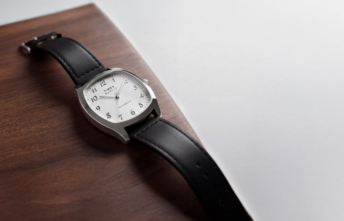 TW2Y84300 Marlin® Chatham Automatic watch with a silver-tone case, white dial and black leather strap resting on a wooden surface.