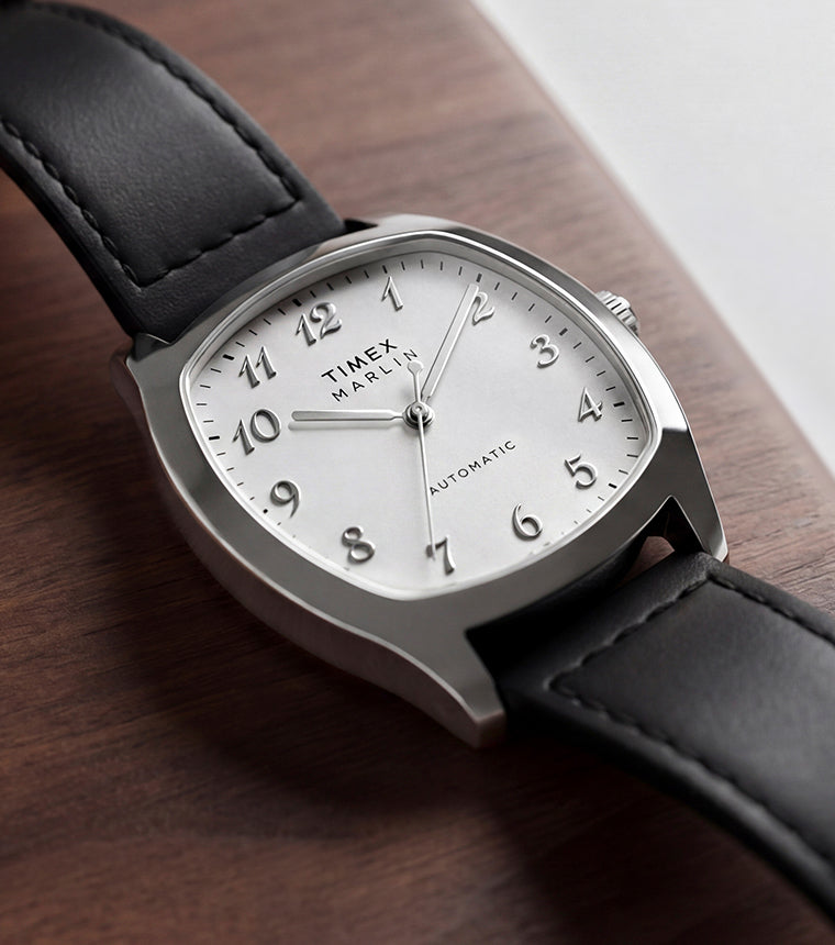 TW2Y84300 Marlin® Chatham Automatic watch with a silver-tone case, white dial and black leather strap resting on a wooden surface.