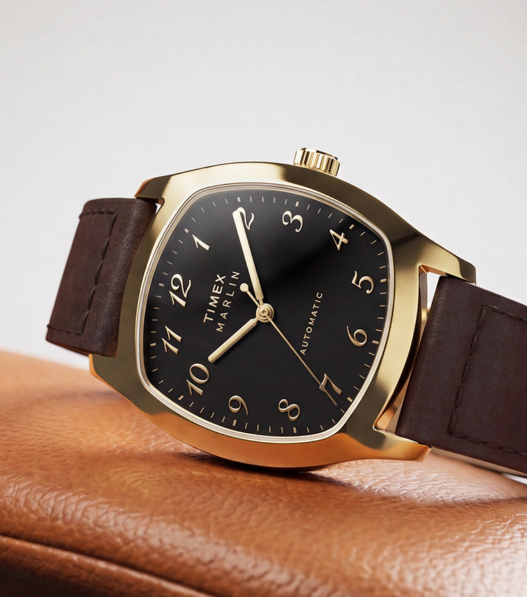 TW2Y84500 Marlin® Chatham Automatic watch with a gold-tone case, black dial and brown leather strap resting on a tan leather chair.