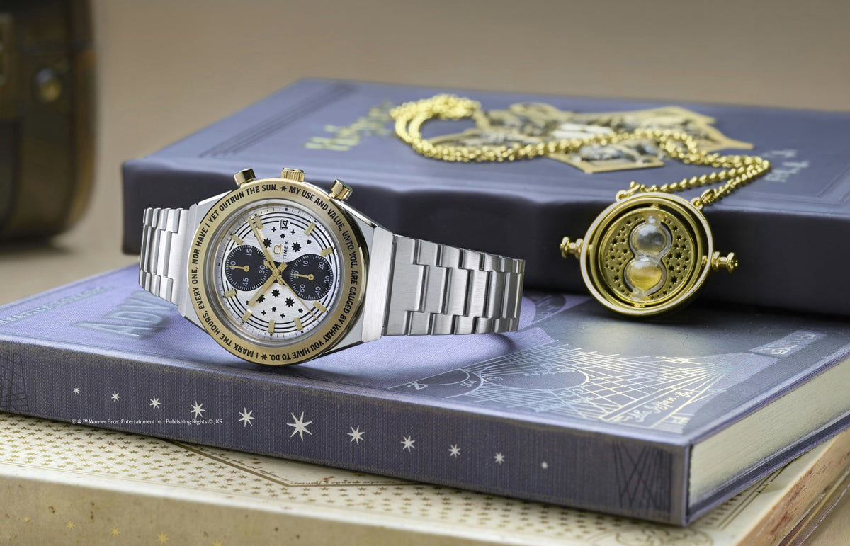 TW2Y75900 Silver-tone chronograph watch with a star-patterned dial and gold-tone bezel displayed beside a gold Time-Turner pendant on a book.