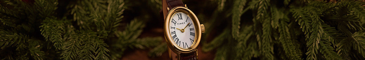 Holiday Favorites - Timex watch with an oval gold-tone case, white Roman numeral dial and brown leather strap among evergreen branches.