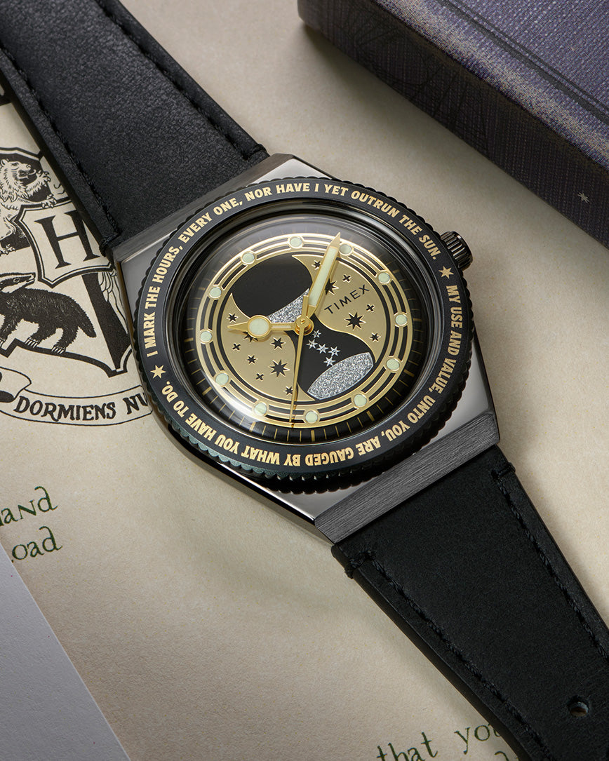 TW2Y76000 Harry Potter x Q Timex Time Turner watch with a black leather strap, gold-tone hands, and a star-patterned dial, resting on illustrated pages beside a book.