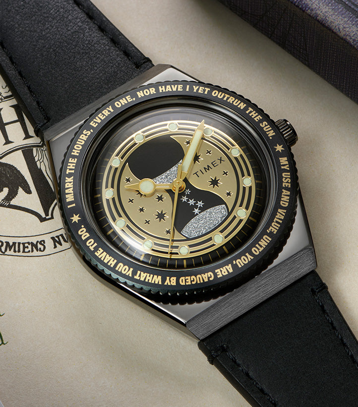 TW2Y76000 Harry Potter x Q Timex Time Turner watch with a black leather strap, gold-tone hands, and a star-patterned dial, resting on illustrated pages beside a book.