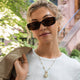 TW6A17700 Model wearing a gold-tone Cavatina® Mini watch necklace over a white top, with sunglasses and a beige jacket over one shoulder.