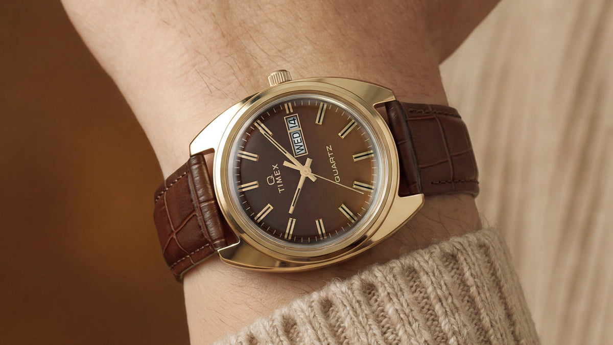 TW2Y87200 Timex 1978 Day Date Fumé Reissue with a brown gradient dial and gold-tone case worn on a wrist beside a beige knit cuff.