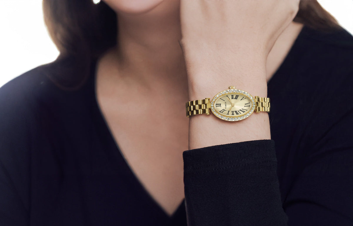 TW2Y90100 Gold-tone Timex Cavatina® Crystals watch with a champagne oval dial and crystal-lined bezel, worn on a raised wrist beside a black sleeve.