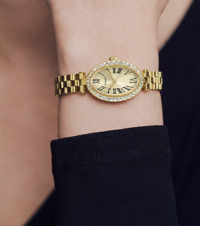 TW2Y90100 Gold-tone Timex Cavatina® Crystals watch with a champagne oval dial and crystal-lined bezel, worn on a raised wrist beside a black sleeve.