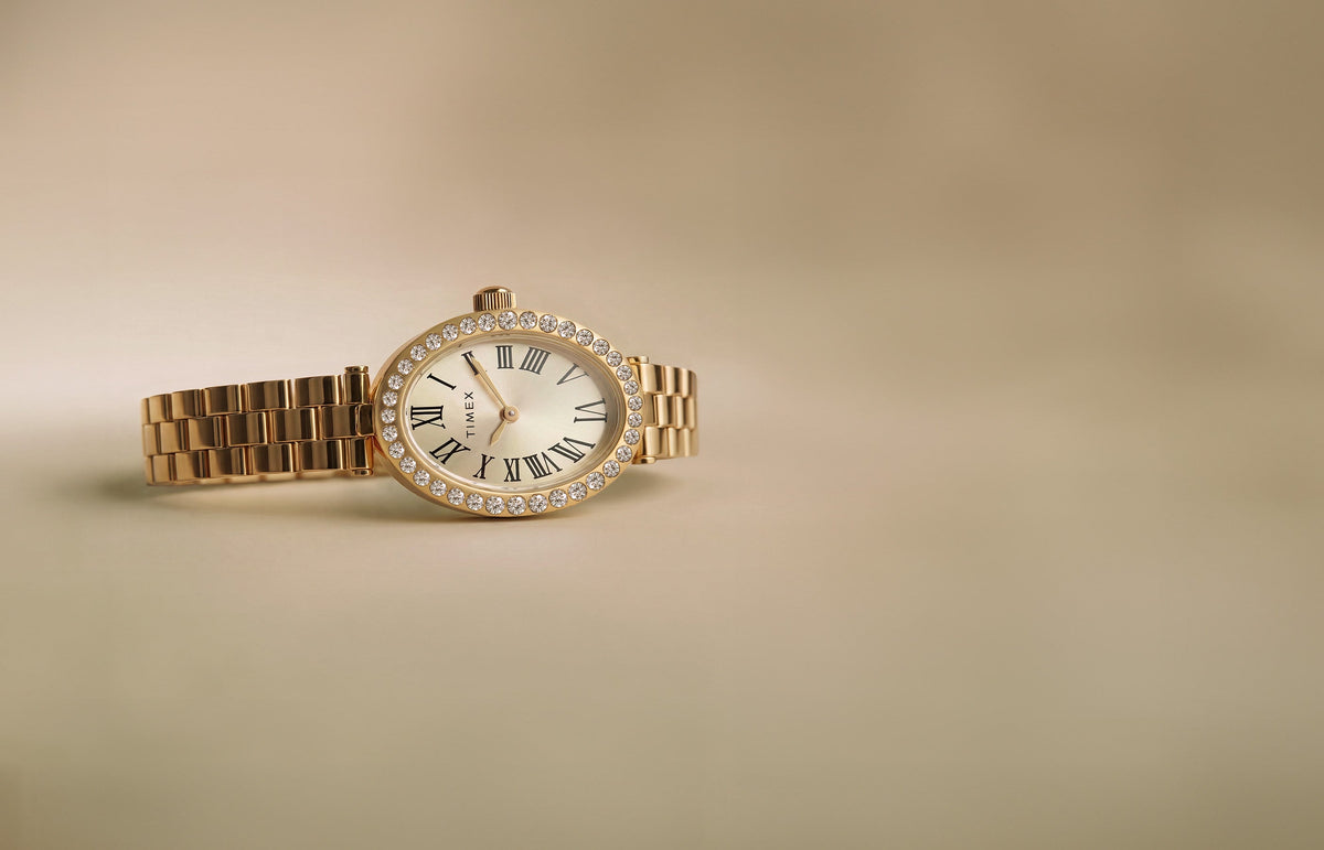 TW2Y90100 Timex Cavatina® Crystals watch with a gold-tone bracelet, champagne oval dial, and crystal-lined bezel on a beige background.