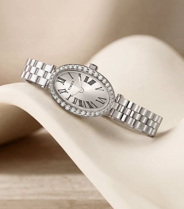 TW2Y90000 Timex Cavatina® Crystals watch with an oval Roman numeral dial, crystal-lined bezel, and silver-tone bracelet, resting on a curved ivory display.