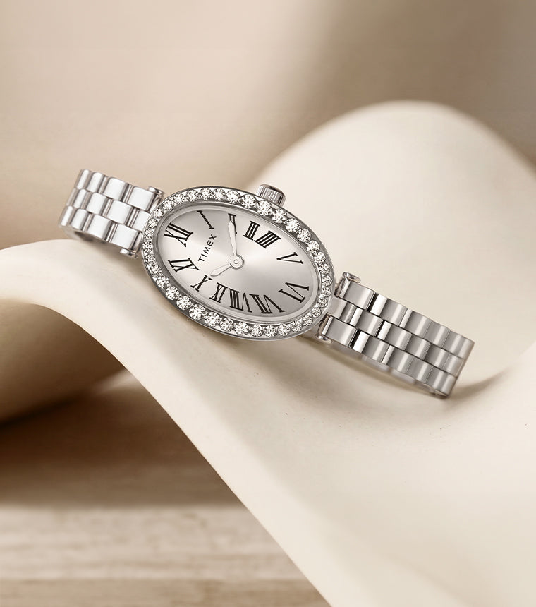 TW2Y90000 Timex Cavatina® Crystals watch with an oval Roman numeral dial, crystal-lined bezel, and silver-tone bracelet, resting on a curved ivory display.
