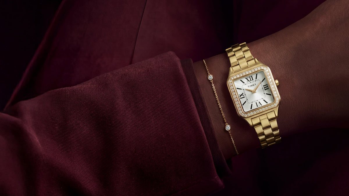 TW2Y90500 Gold-tone Timex Addison watch with a crystal-lined bezel and white Roman numeral dial, styled with a delicate bracelet and burgundy outfit.