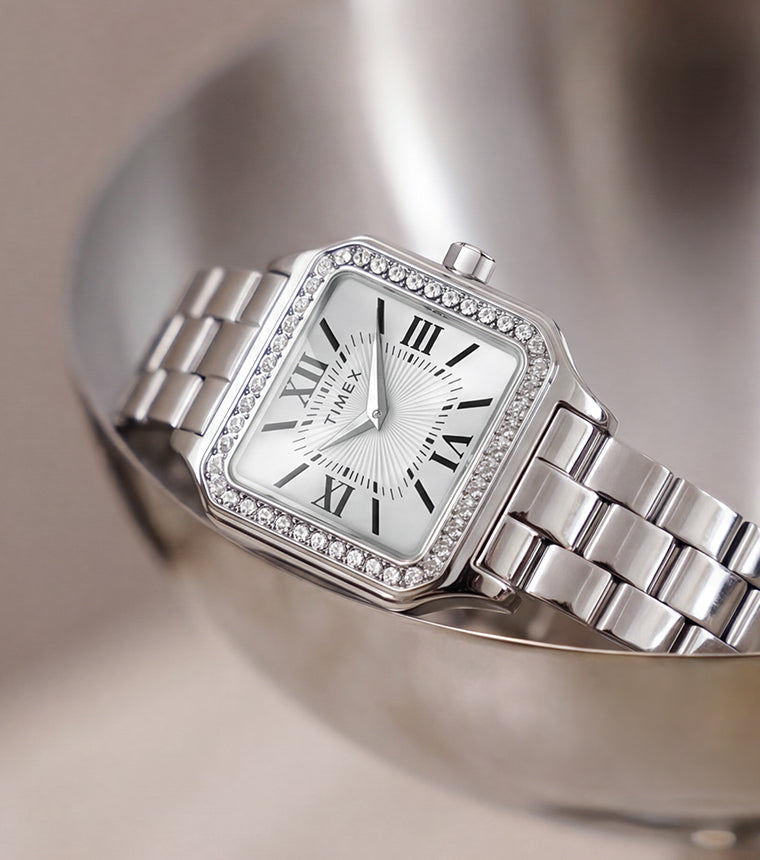 TW2Y90300 Timex Addison watch with a silver-tone bracelet, white Roman numeral dial, and crystal-lined bezel, resting in a metal dish.