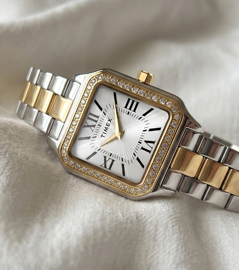 TW2Y90400 Timex Addison watch with a silver-and-gold-tone bracelet, white Roman numeral dial, and crystal-lined bezel, resting on ivory fabric.