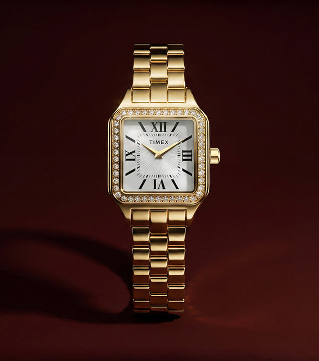 TW2Y90500 Gold-tone Timex Addison watch with a white Roman numeral dial, crystal-lined rectangular bezel, and link bracelet against a dark burgundy background.