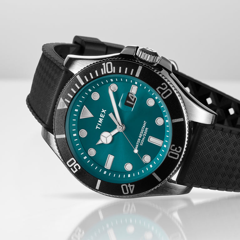 A close-up angled shot of the Timex Harborside Coast watch resting on a reflective white surface. The watch features a vivid teal blue dial with bold white hour markers, silver-toned hands, and a magnified date window at the 3 o’clock position. A unidirectional black bezel with white markings encircles the face, adding a classic dive watch aesthetic. The watch is paired with a textured black silicone strap and brushed stainless steel case, exuding a sporty and durable look.