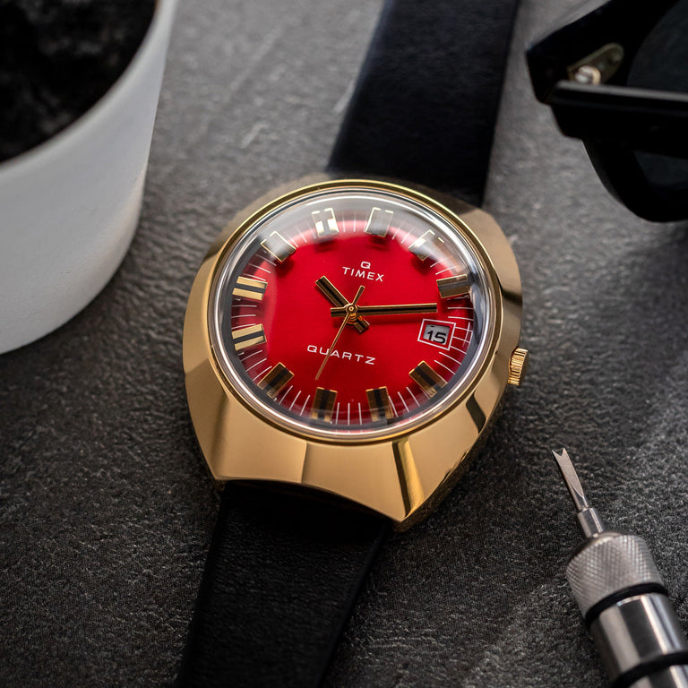 Timex Q Reissue watch with a bold red dial, gold-tone case, black leather strap, and date window, displayed on a textured surface with watchmaking tools and accessories.