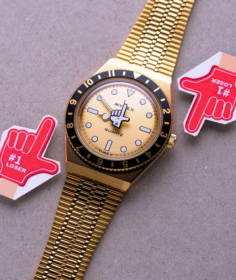 Gold seconde/seconde watch on a grey background with red hand icons either side