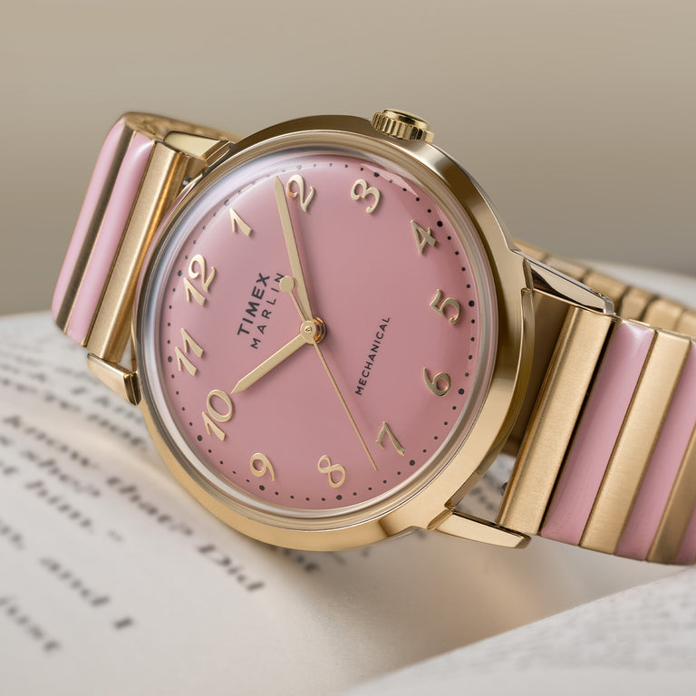 Timex women’s Marlin watch with a soft pink dial, gold-tone case, Arabic numerals, and a gold bracelet with pink enamel accents, shown in a close-up product shot.