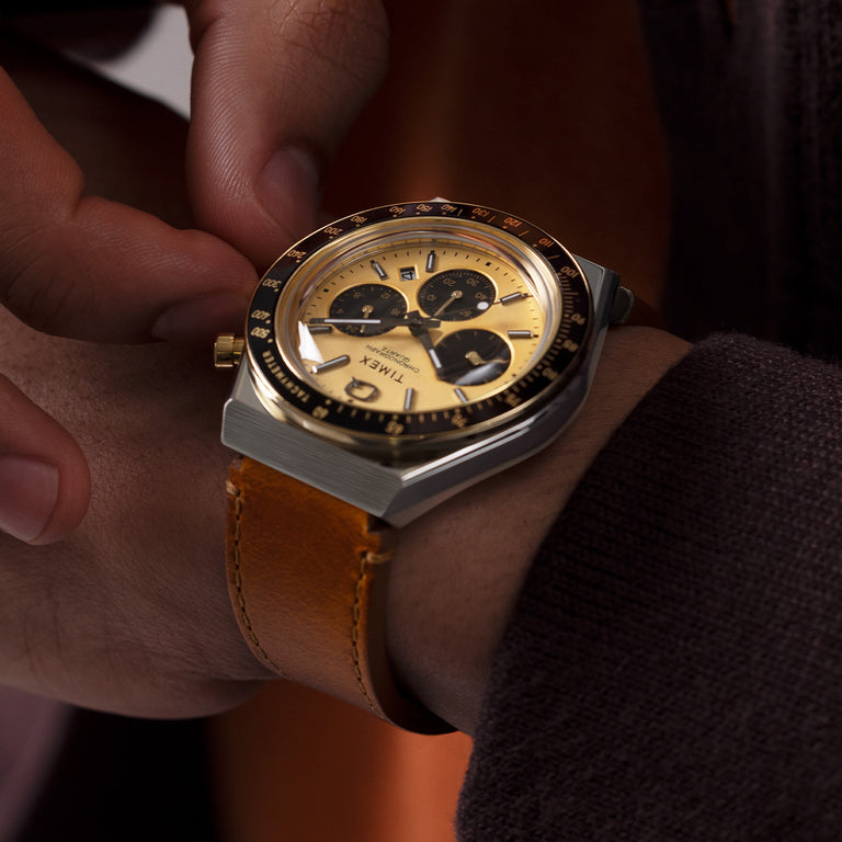A close-up shot of a Q Timex Chronograph watch on a person’s wrist. The watch features a brushed stainless steel case with a gold-tone dial, three black sub-dials, and a date window between the 4 and 5 o’clock positions. It has a black tachymeter bezel and luminous hands with polished hour markers. The timepiece is paired with a rich brown leather strap, and the wearer is adjusting the crown with one hand, dressed in a dark knit sweater and warm-toned layers.