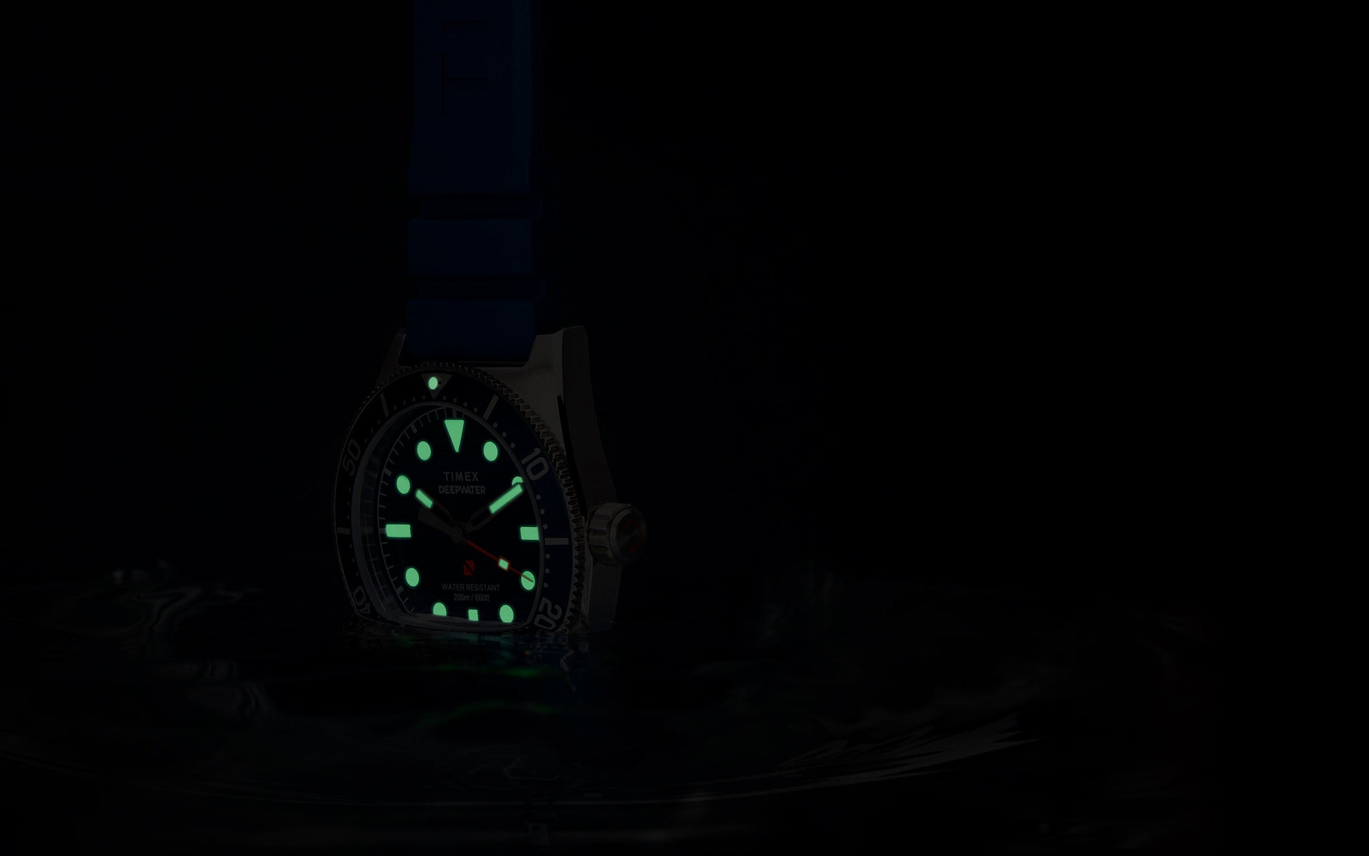The Timex Deepwater Reef 200 glowing in low light. Its hands, hour markers, and bezel pip emit a vivid green luminescence while the red seconds hand remains faintly visible. The watch floats just above water, enhancing its dive functionality.