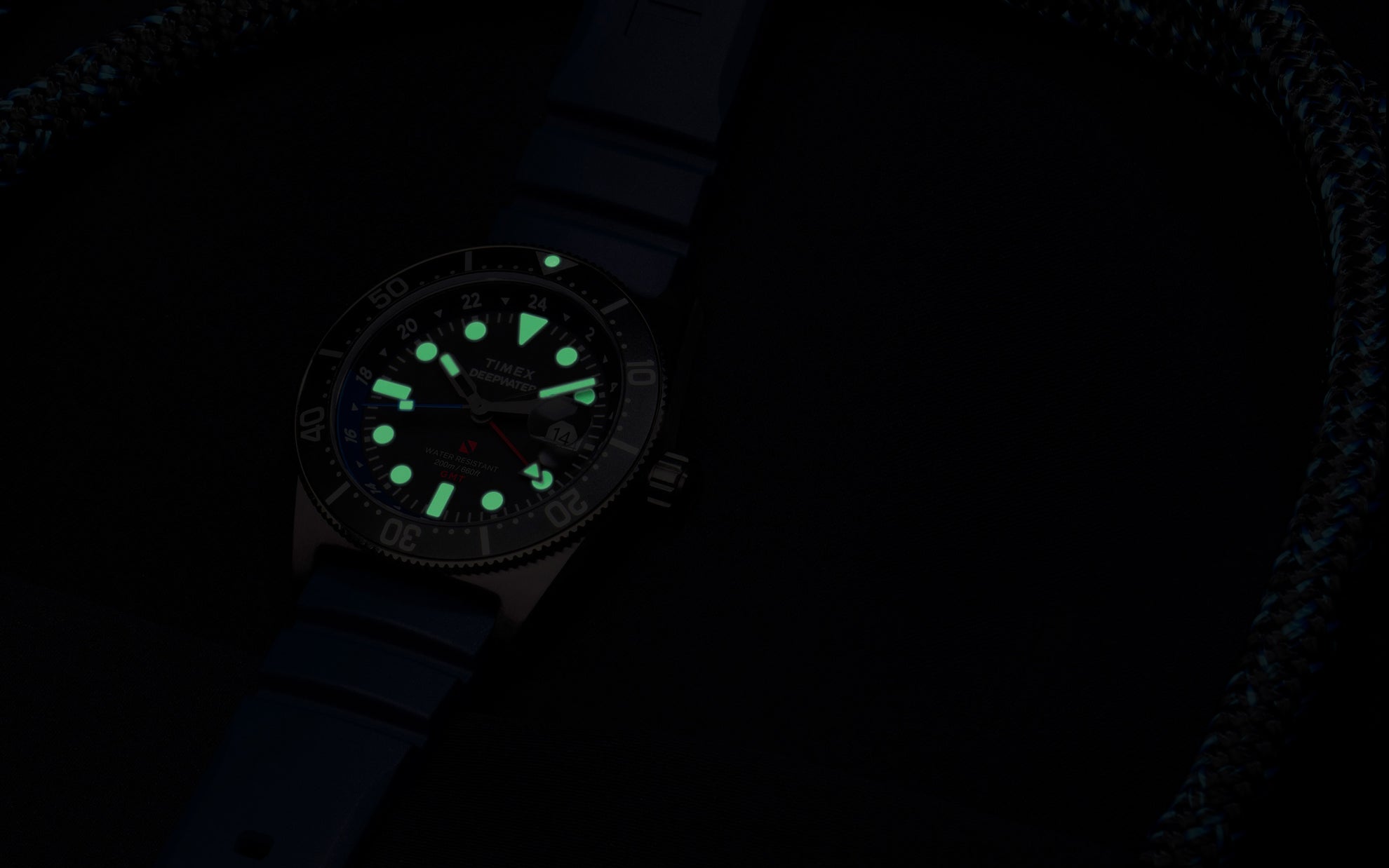 The same Timex Deepwater Reef 200 GMT watch in low light. Its hands, hour markers, and bezel pip emit a soft green glow, showcasing its high underwater visibility.