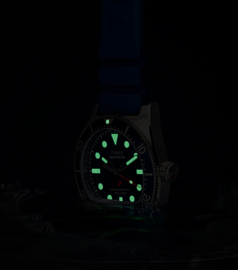 The Timex Deepwater Reef 200 glowing in low light. Its hands, hour markers, and bezel pip emit a vivid green luminescence while the red seconds hand remains faintly visible. The watch floats just above water, enhancing its dive functionality.