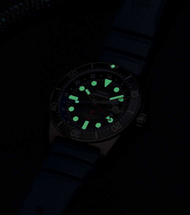 The same Timex Deepwater Reef 200 GMT watch in low light. Its hands, hour markers, and bezel pip emit a soft green glow, showcasing its high underwater visibility.