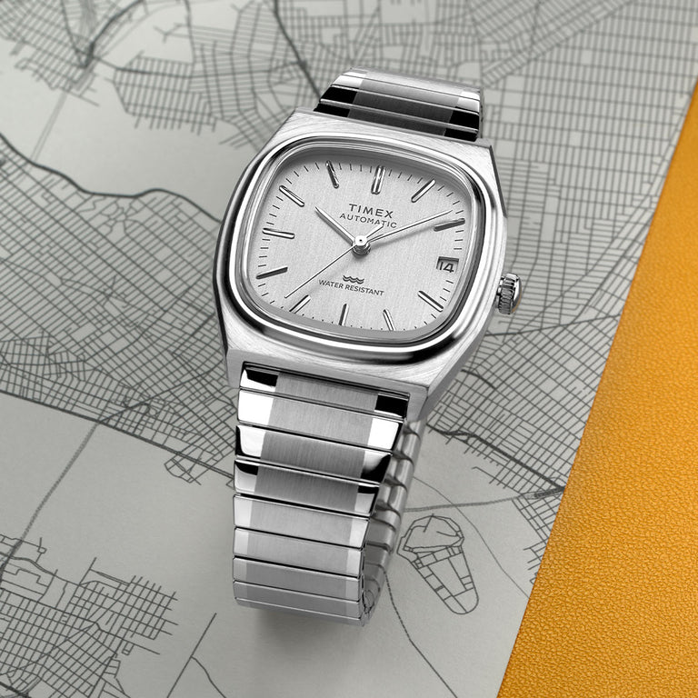 Timex Eline Automatic watch with a silver-tone square cushion case, silver sunray dial, and stainless steel bracelet. The dial features slim hour markers, a date window at 3 o’clock, and water resistance text. The watch is displayed on a city map background with a leather accent.