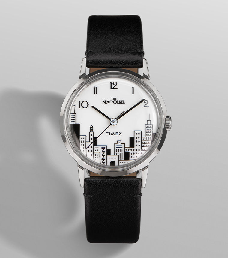 A detailed close-up of the Timex x The New Yorker Marlin® Hand-Wound watch. The dial is white with black line-art illustrations of a New York City skyline at the bottom. The hands are sleek, with a black minute and hour hand, and a silver second hand. The words "The New Yorker" are displayed below the 12 o’clock position, with "Timex" centered near the skyline. The polished stainless-steel case and black leather strap add to its elegant design.