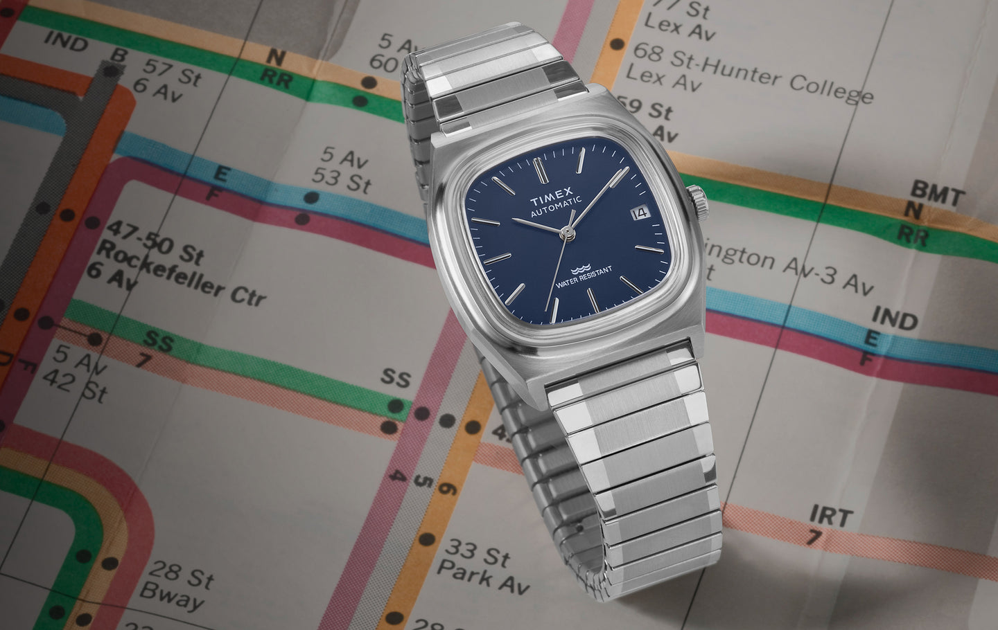 A stainless steel Timex E-Line Reissue watch with a blue dial is displayed against a vintage New York City subway map. The watch features a sleek square case, automatic movement, date display, and a water-resistant design. The subway map in the background highlights iconic NYC locations.